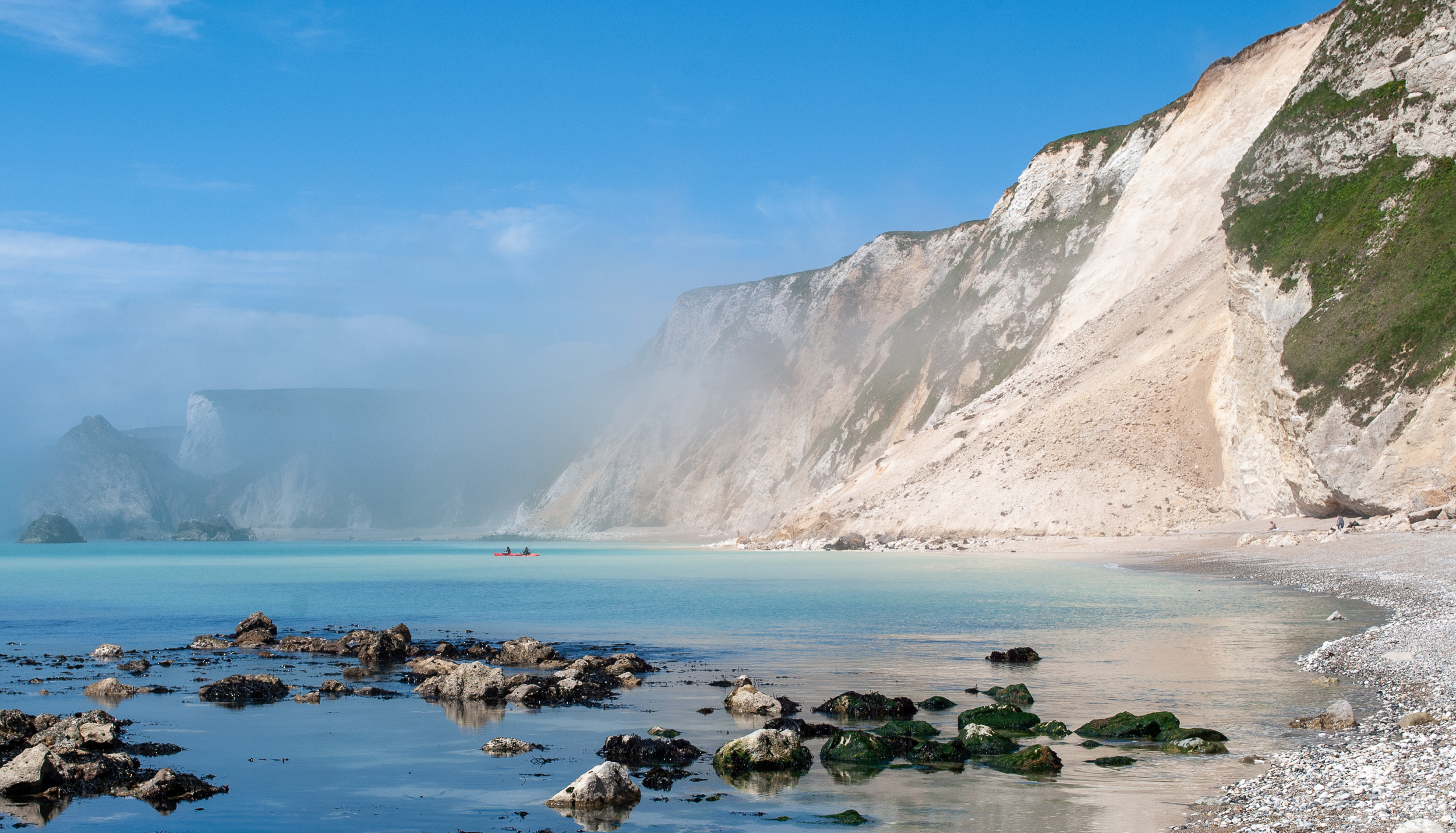 Photo of a landslip in the white cliffs in St Oswald's Bay, near Lulworth Cove in Dorset. The water is milky with chalk and two tiny canoeists are dwarfed by the cliffs and the landslip